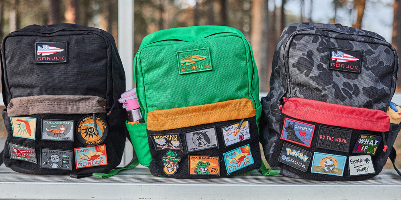 Three GORUCK backpacks with colorful patches on a bench, surrounded by trees.