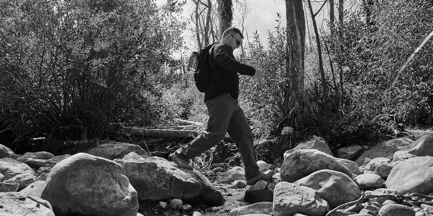 Man hiking through rocky terrain in a forested area, wearing a backpack. Black and white image.