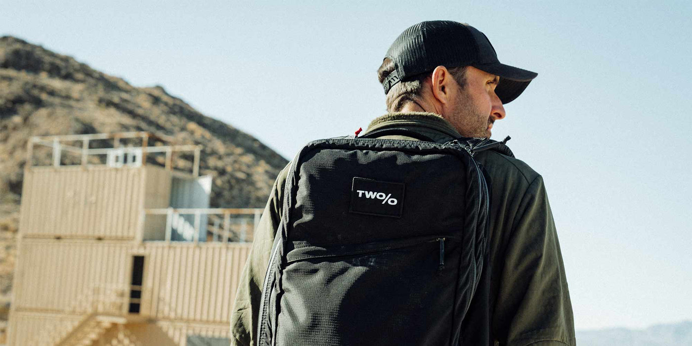 Man wearing a hat and backpack stands outdoors near a building and hill.