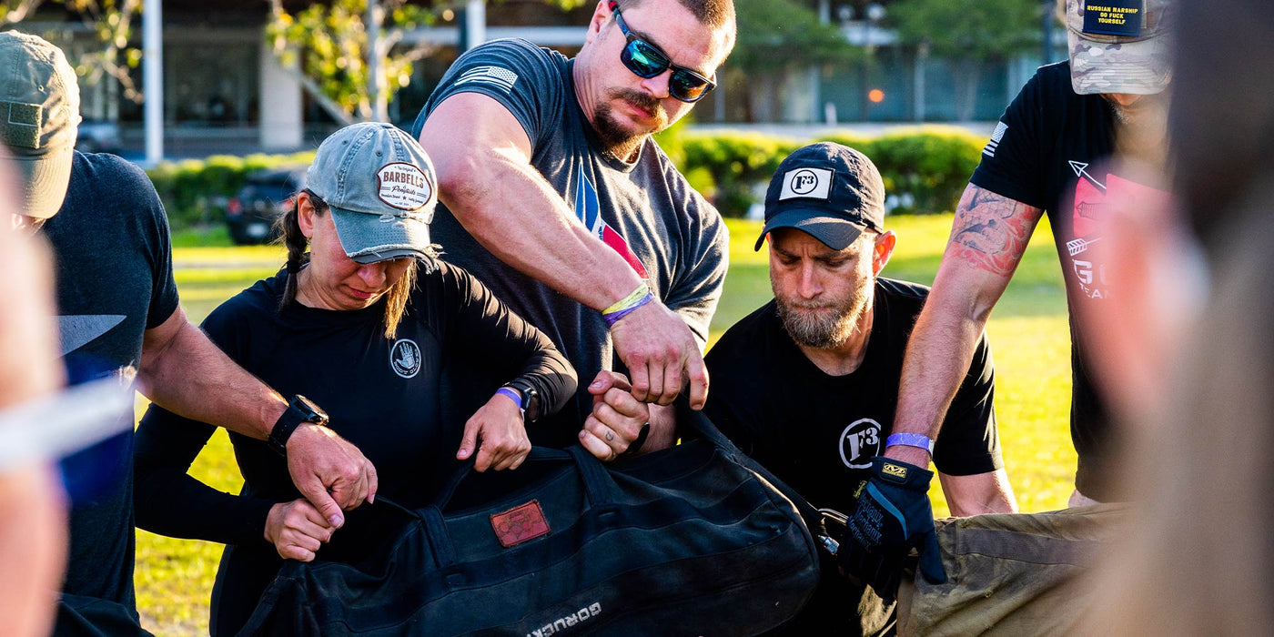 A group of people working together outdoors, lifting a large sandbag during a team-building event.