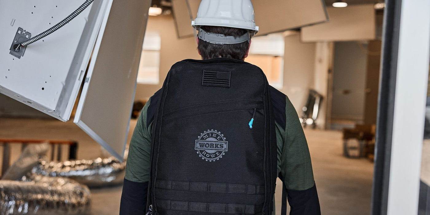 Person wearing a black backpack with "Works" logo, in an indoor setting with construction materials nearby.