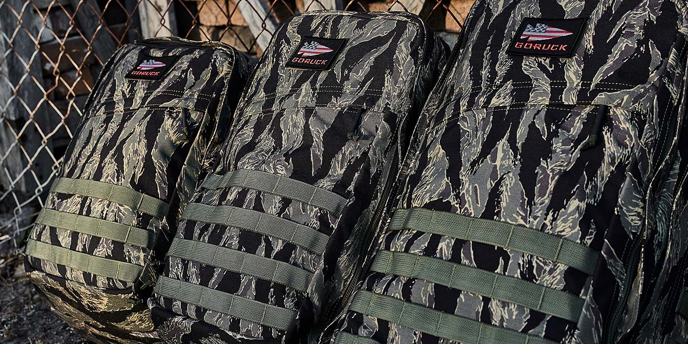 Three camo backpacks with MOLLE webbing lined up against a chain-link fence.