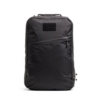 The GR1 USA - X-PAC is a sleek black rucksack featuring a minimal design with a zippered front pocket and small top patch. Crafted from waterproof X-PAC material, it combines style and durability for everyday use.