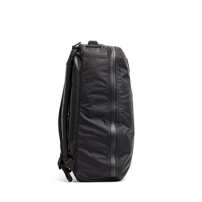 Side view of the GR1 USA - X-PAC, a sleek black rucksack with multiple zippers, made from X-PAC material and standing upright on a white background.