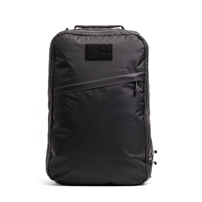 The GORUCK GR1 USA - X-PAC backpack boasts a sleek black design with AquaGuard zippers and a front zipper pocket, complemented by a durable X-PAC construction and a convenient top handle, all set against a pristine white backdrop.