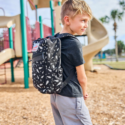 A young boy stands on a playground, wearing a black T-shirt, gray shorts, and the stylish GORUCK KR1 - Spearhead Splatter kids rucksack. The playground slide and equipment are in the background on a sunny day.