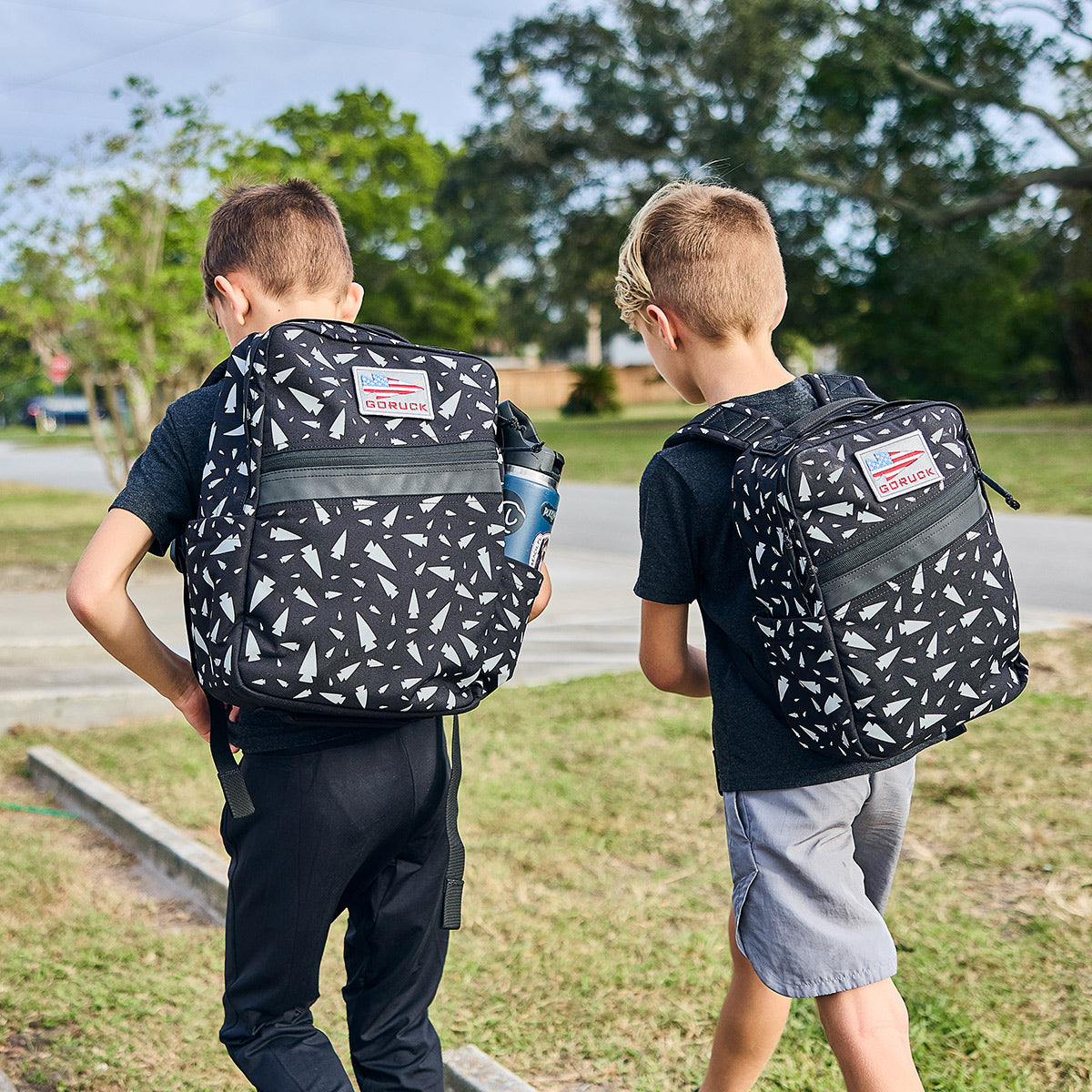 Two children equipped with GORUCK's KR1 - Spearhead Splatter book bags, featuring black and white geometric patterns, walk outdoors on a sunny day. One of them is carrying a water bottle. They are surrounded by trees and grass, with a sidewalk visible in the background.