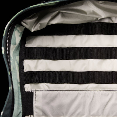 The image showcases the interior of the GR1 USA x Gear Patrol, a limited-edition camouflage backpack by GORUCK. It features a gray lining with black horizontal straps for organization, seamlessly integrated into the design, reminiscent of variegated elephant ear camo.