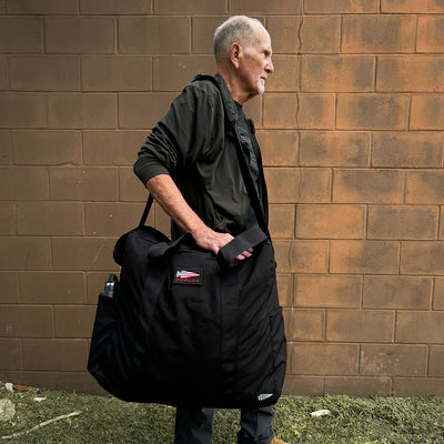 A man with short hair stands against a brown brick wall, carrying a GORUCK Kit Bag - 84L, which features a large black design and a USA flag patch. He wears a dark jacket and pants, and from the bag's pocket emerges a silver water bottle.