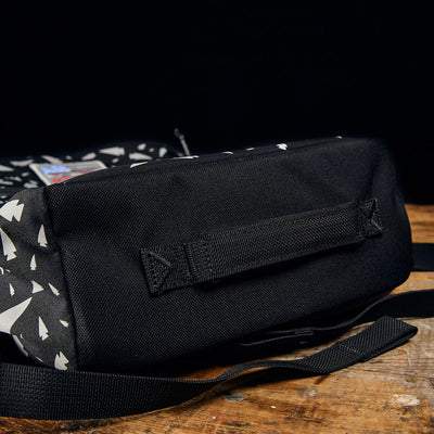 The KR1 - Spearhead Splatter from GORUCK, featuring a striking geometric pattern, rests on a wooden surface against a black background. It comes equipped with a fabric handle on its side and an adjustable shoulder strap, making it ideal for carrying school supplies.