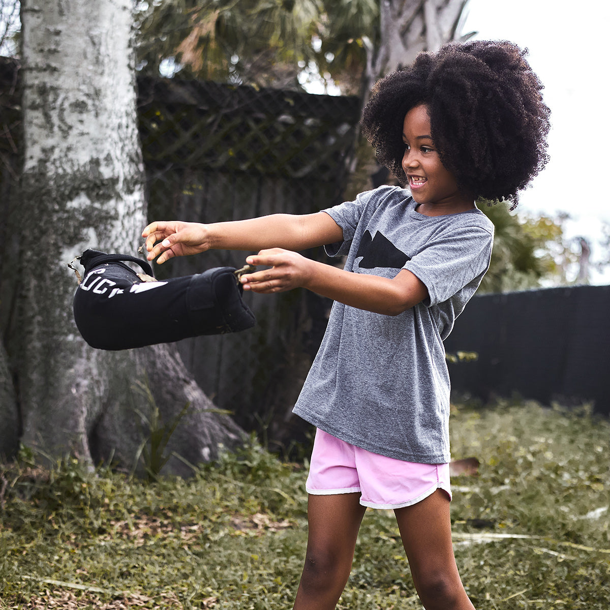 A joyful child with curly hair, dressed in a gray Batman shirt and pink shorts, plays outside wearing a black jacket. They stand before a tree and fence amidst verdant grass and foliage, participating in GORUCK's Elementary School Ruck Training Program that emphasizes enjoyable, functional movements for development.