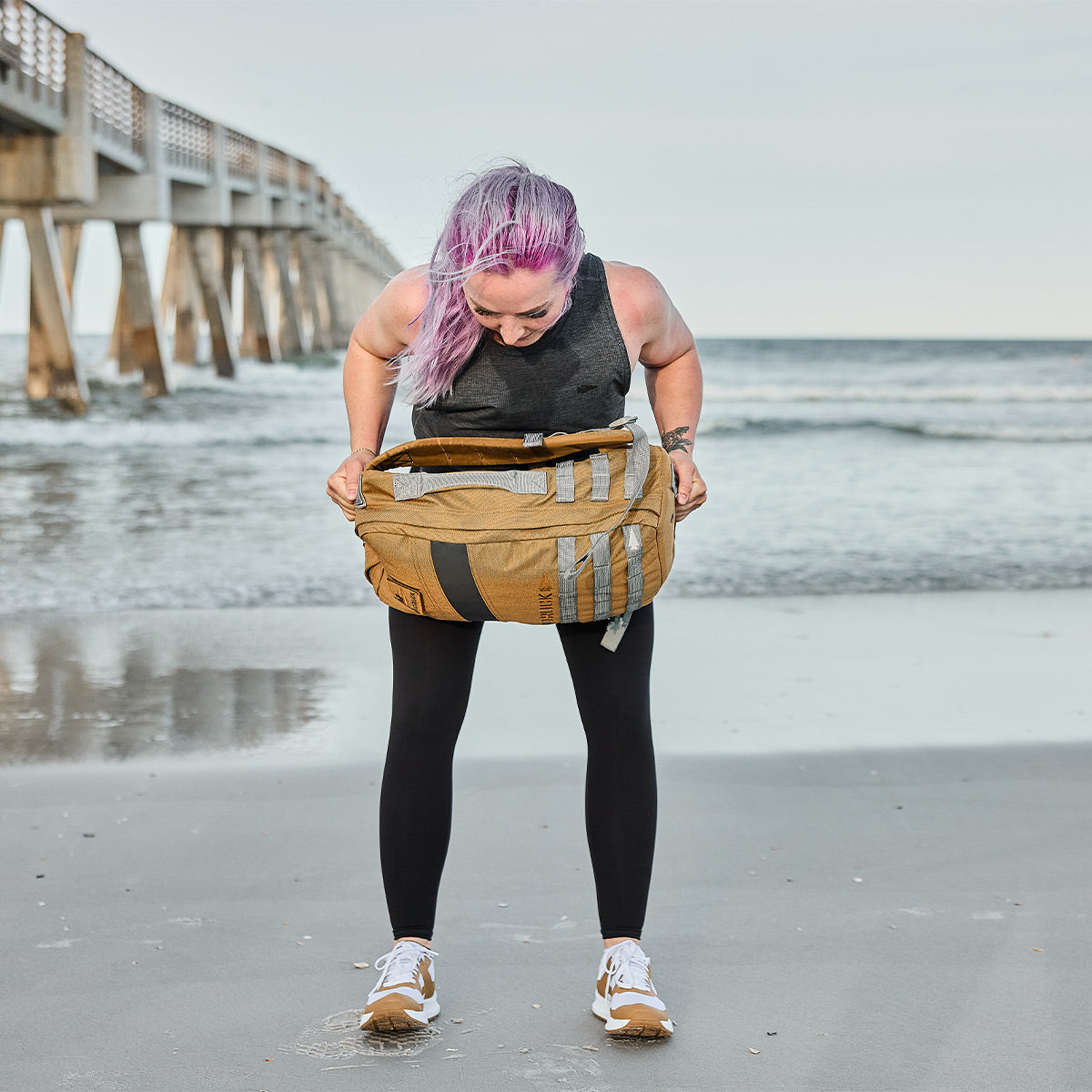 A person with purple hair stands on a sandy beach, checking their large brown and gray backpack. They're wearing a black top and leggings, with Women's Rough Runner - Speed Tan shoes by GORUCK. A pier extends into the ocean in the background.