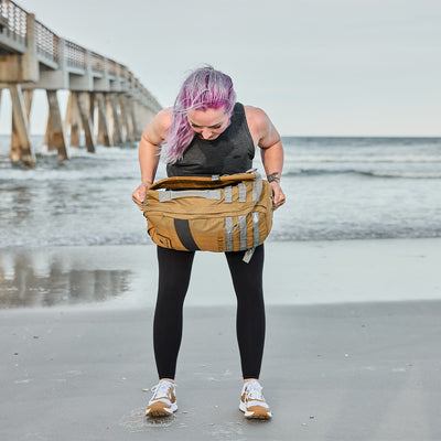 A person with purple hair stands on a sandy beach, checking their large brown and gray backpack. They're wearing a black top and leggings, with Women's Rough Runner - Speed Tan shoes by GORUCK. A pier extends into the ocean in the background.