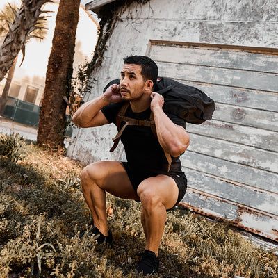 A man squats outdoors with a backpack, wearing GORUCK's Men’s Ranger Panties - ToughStretch, in front of a weathered garage and palm tree.