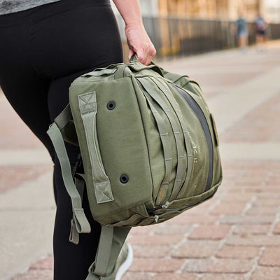 Carrying a Rucker 4.0 backpack by a strap and featuring a Ruck Plate®, a person strolls confidently along the paved path.