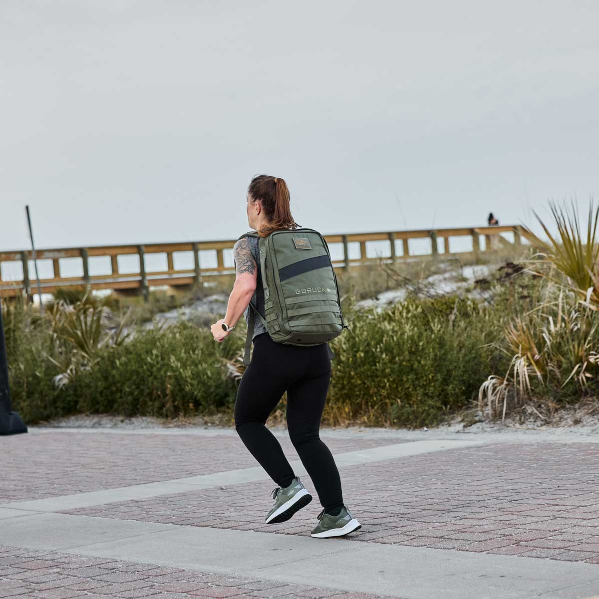 Someone carrying a Rucker 4.0 strolls along a pathway near the boardwalk and grassy area, possibly using it for rucking with added weight.