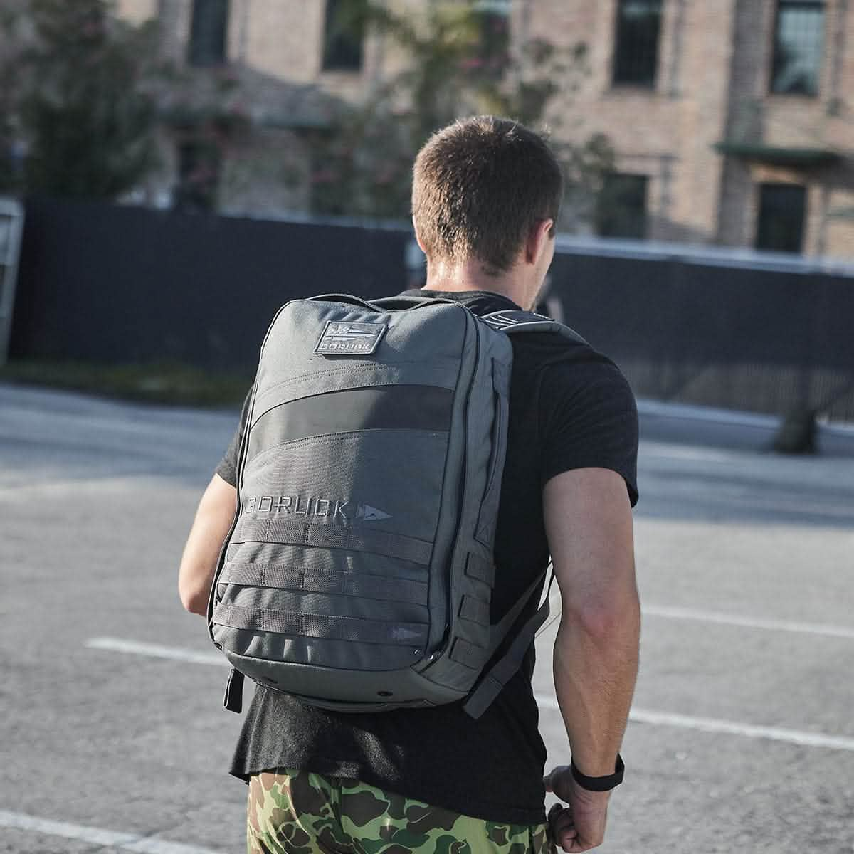 A person wearing a black T-shirt and camouflage shorts is outdoors, facing away from the camera. They are carrying a large gray Rucker 4.0 rucksack from GORUCK on their back. The background showcases a brick building and a fenced area, perfect for rucking enthusiasts.