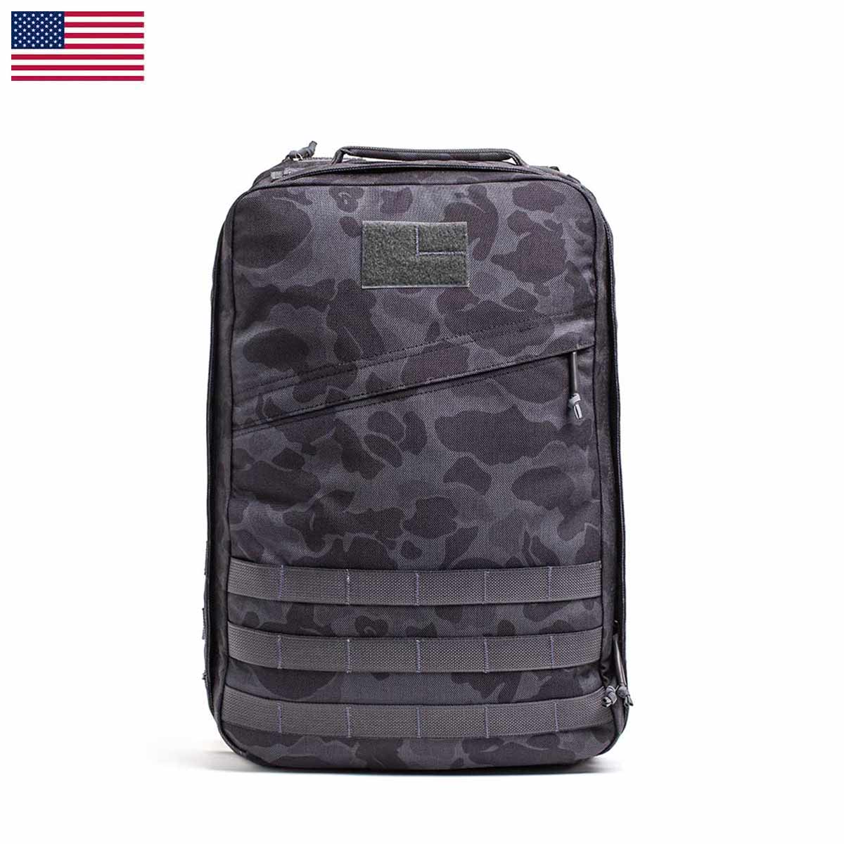 The GR1 USA - Cordura (The Original Ruck), inspired by Special Forces gear, is a gray camo backpack featuring multiple zippers and straps, proudly adorned with a small American flag in the corner.