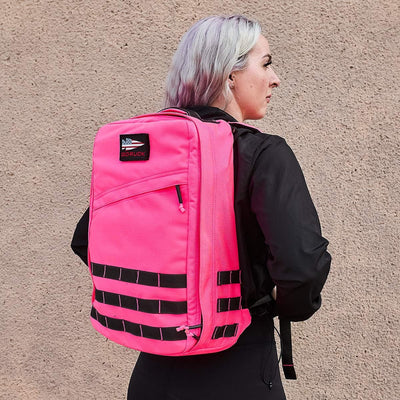 A person with light hair is wearing the GR1 USA - Hot Pink backpack by GORUCK, showcasing a sense of toughness as they stand against a beige textured wall. This performance-focused backpack, featuring multiple compartments and a small logo on the top flap, complements their black jacket.