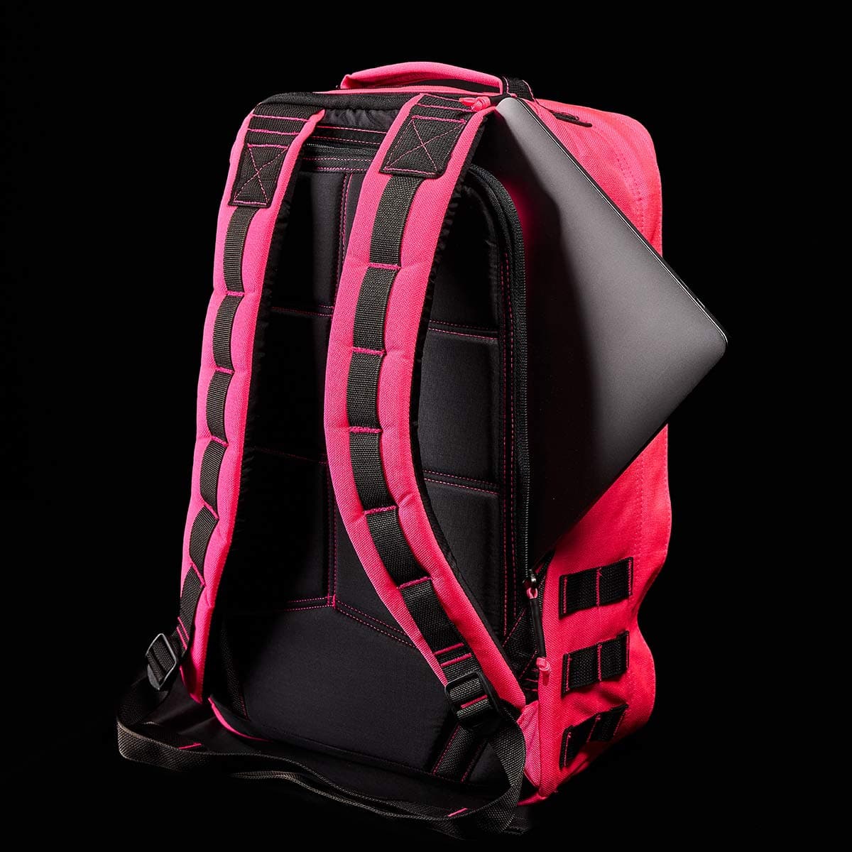 The GR1 USA - Hot Pink backpack by GORUCK is featured against a black backdrop, showcasing its vibrant pink design and black padded straps that resonate with the durability of Special Forces equipment. Peeking from within is a laptop, highlighting its practicality for tech transportation.