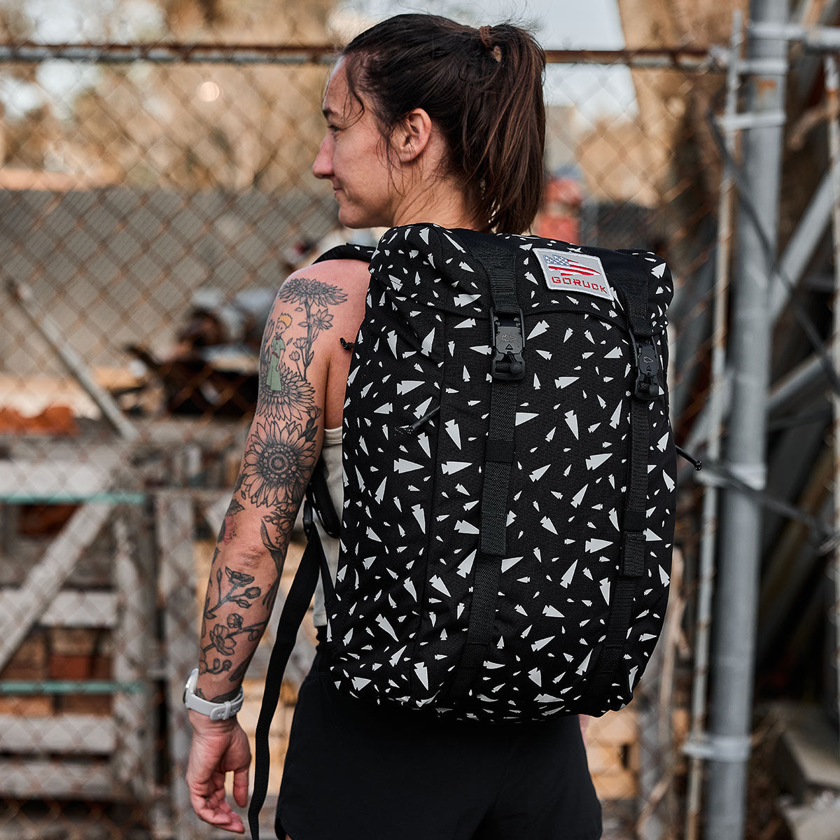 A person with a tattooed arm carries the GORUCK M23 - Cordura - Spearhead Shrapnel rucksack, featuring durable materials and a geometric pattern. They are outdoors, standing near a chain-link fence and wearing a sleeveless top.