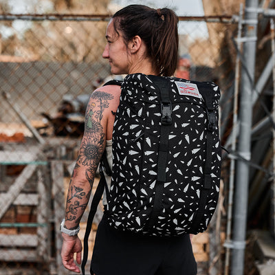 A person with a tattooed arm carries the GORUCK M23 - Cordura - Spearhead Shrapnel rucksack, featuring durable materials and a geometric pattern. They are outdoors, standing near a chain-link fence and wearing a sleeveless top.