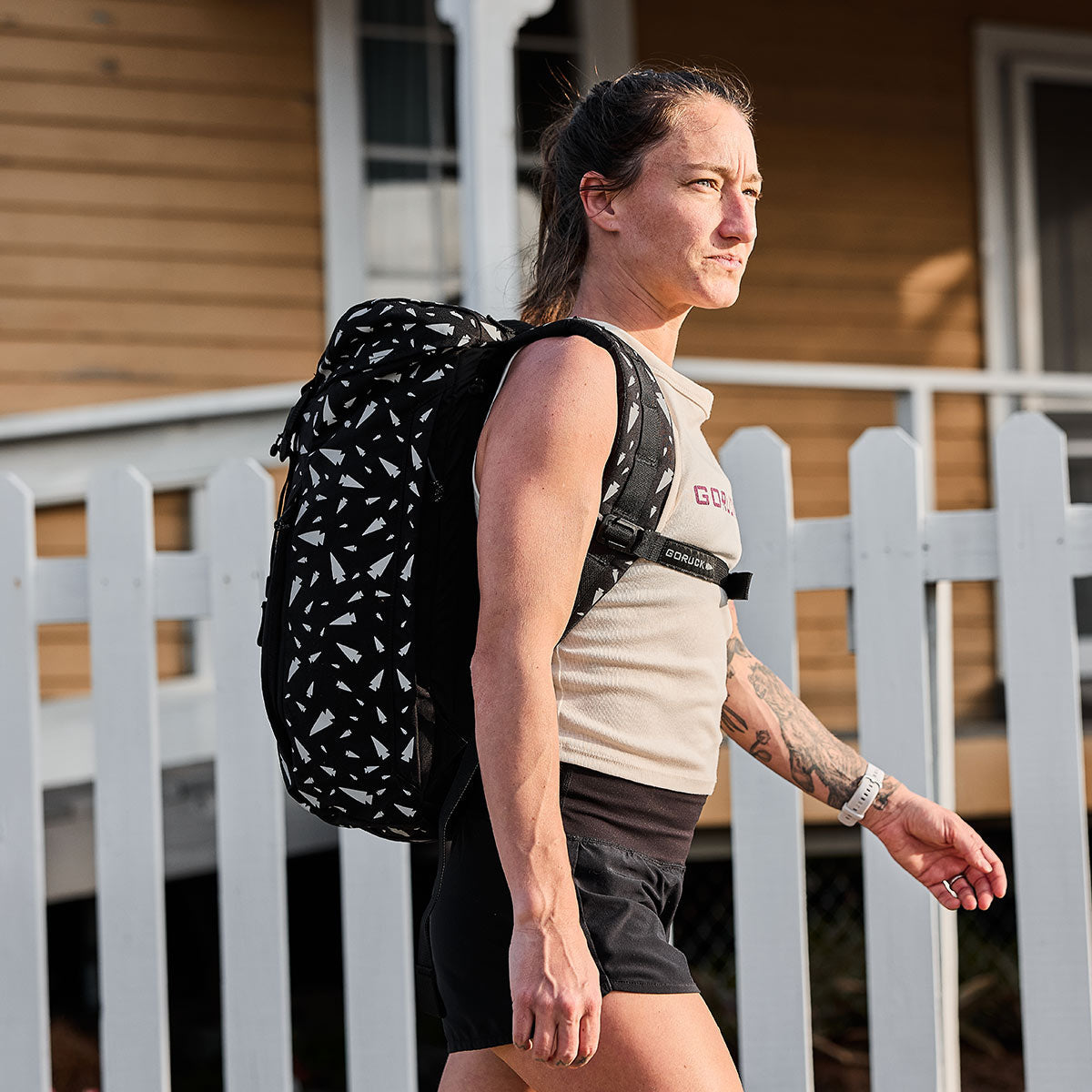 A person with long hair tied back, dressed in a sleeveless shirt and shorts, walks beside a white picket fence. They carry the M23 - Cordura - Spearhead Shrapnel rucksack from GORUCK, featuring a black and white pattern made from durable materials. The background showcases a wooden house with windows.