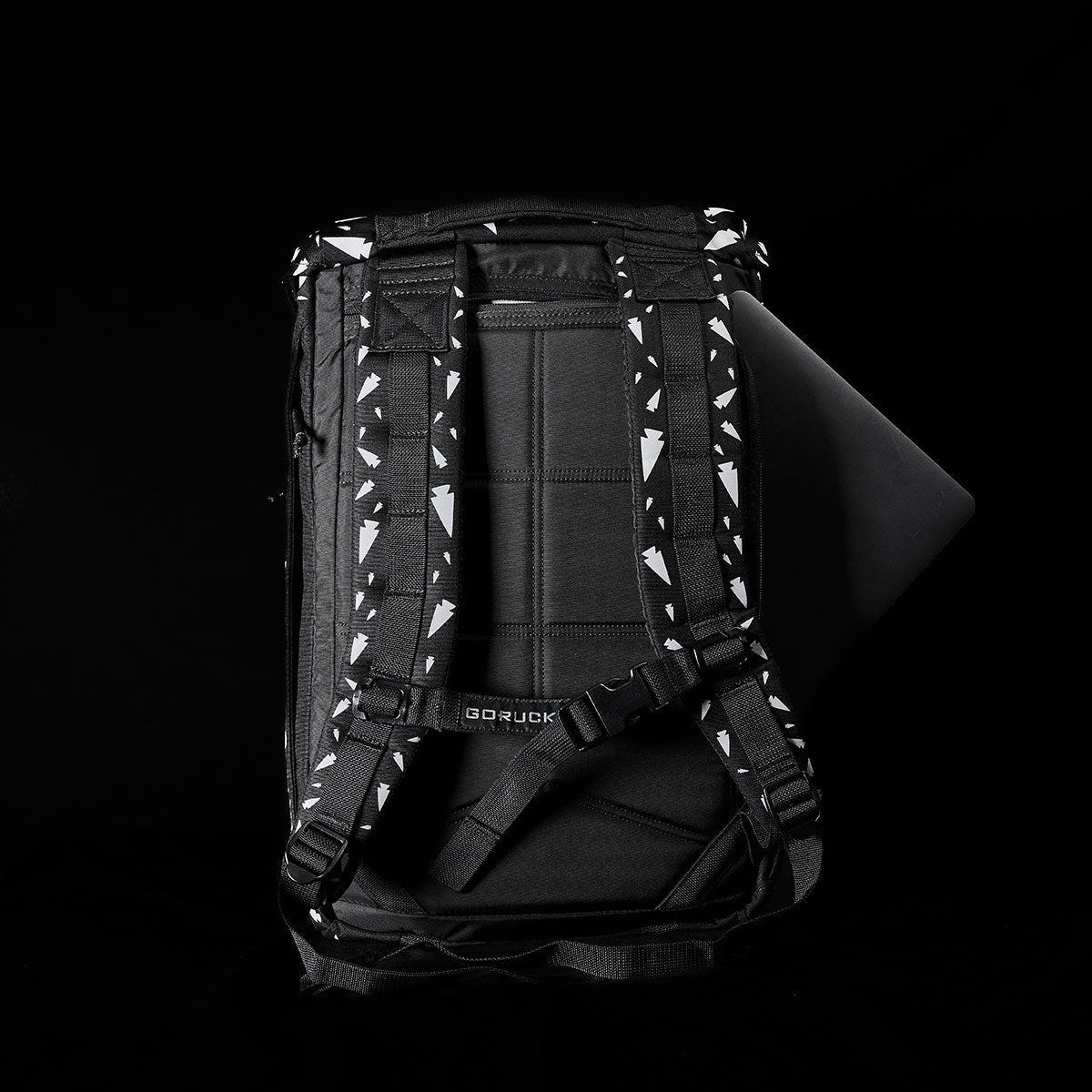 The GORUCK M23 - Cordura - Spearhead Shrapnel, a stylish black rucksack with patterned shoulder straps, is opened halfway to reveal a laptop inside. Crafted from durable materials, this accessory stands out against the solid black background, showcasing its sleek design.