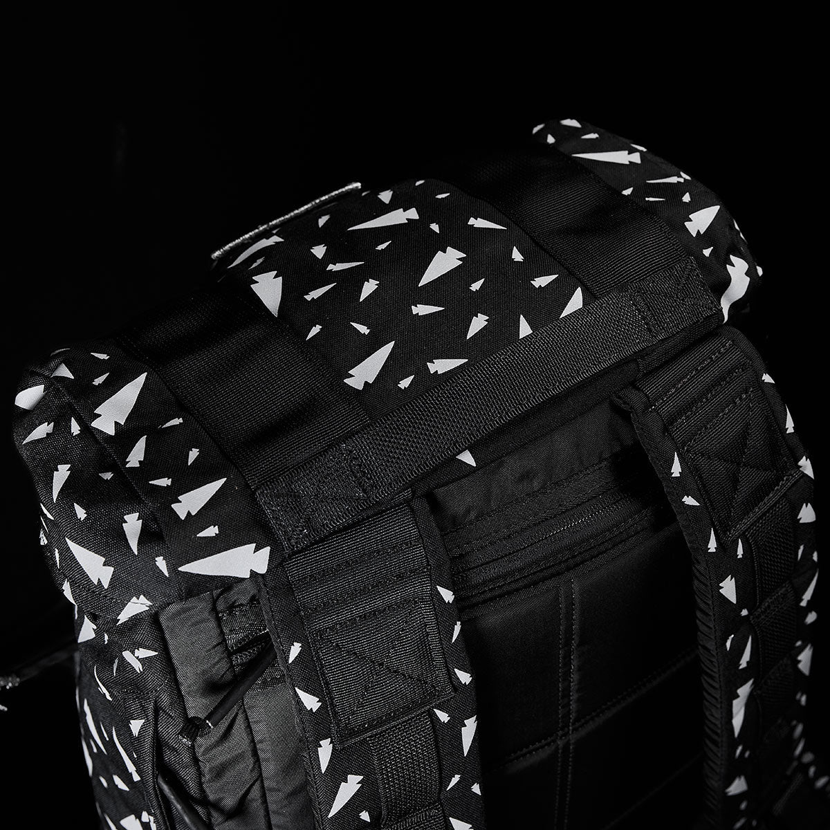 Close-up of the GORUCK M23 - Cordura - Spearhead Shrapnel rucksack showcasing a black and white geometric pattern. The design features white triangles scattered across the durable fabric. This backpack has padded shoulder straps and a top handle, reminiscent of an M1945 Field Pack, set against a dark background.