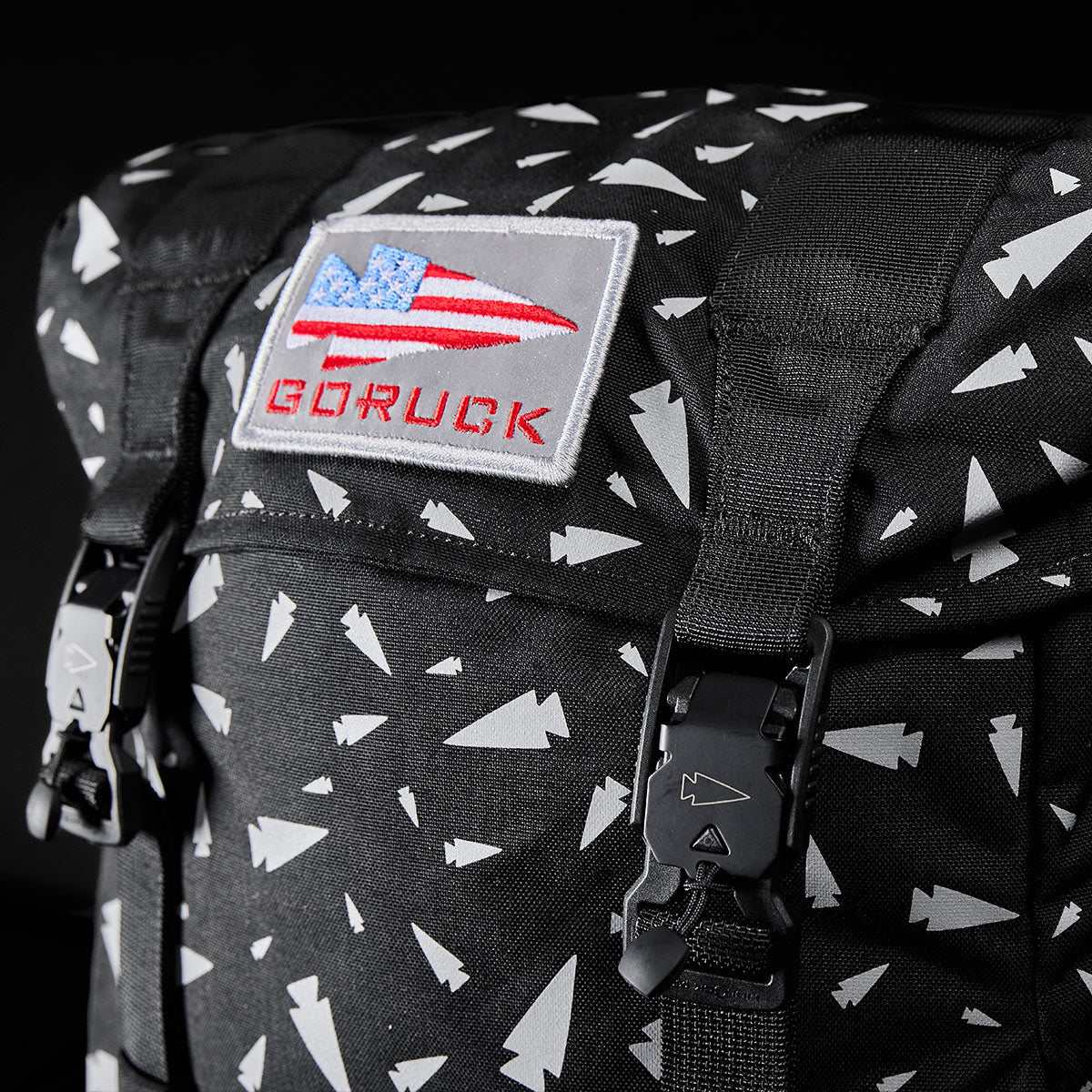 Close-up of the GORUCK M23 Spearhead Shrapnel rucksack featuring a black design with white geometric patterns. The front flap displays a bold patch of the American flag along with the "GORUCK" text. This bag is crafted from durable Cordura material and includes black buckles to secure the top.