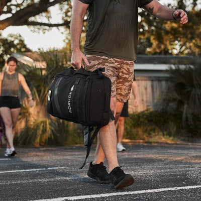 A man in camo shorts with a GORUCK Rucker 4.0 backpack strides confidently outdoors; trees and fellow adventurers frame the scene, likely engaged in rucking.