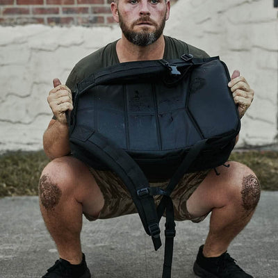 A man squats with a heavy GORUCK Rucker 4.0 backpack, knees muddy, wearing a green shirt and camo shorts on a concrete surface.