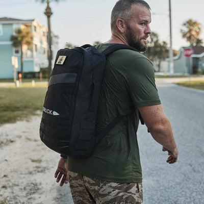 A man rucking outdoors with his large black Rucker 4.0 backpack from GORUCK, sporting a green shirt and camo shorts, enjoys the sunny day.