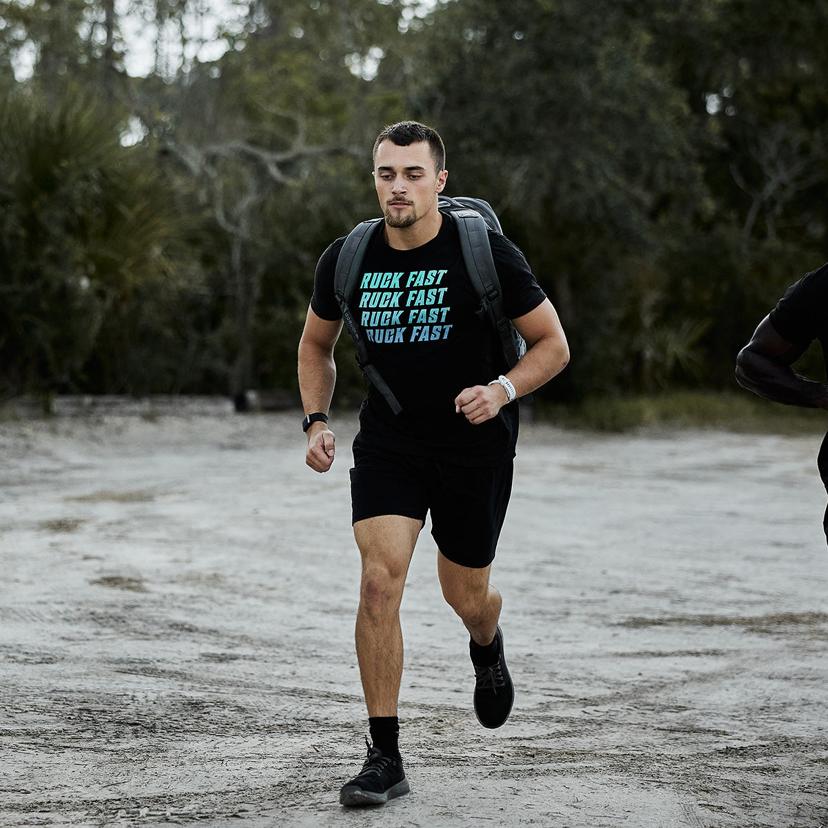 A person is rucking outdoors along a dirt path, clad in a "RUCK FAST" T-shirt, shorts, sneakers, and a wristwatch while carrying the Rucker 4.0 backpack from GORUCK. The background displays trees and foliage.