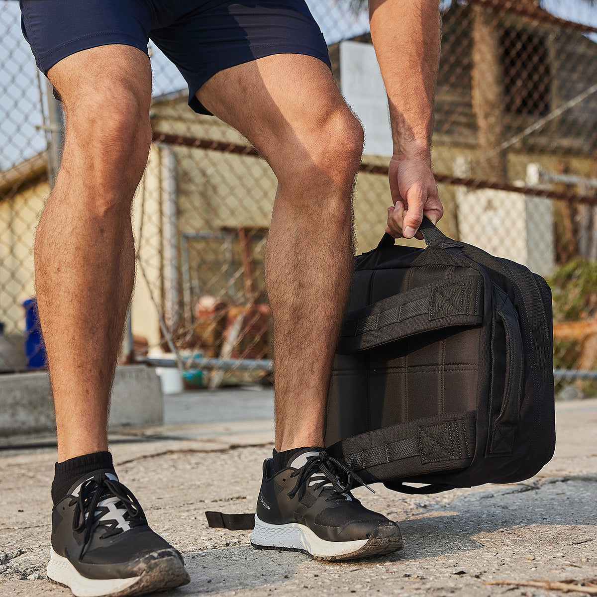 A person wearing black sneakers and navy shorts squats down to pick up a GORUCK Rucker 4.0 rucksack in an outdoor urban setting, framed by a chain-link fence and building in the background.