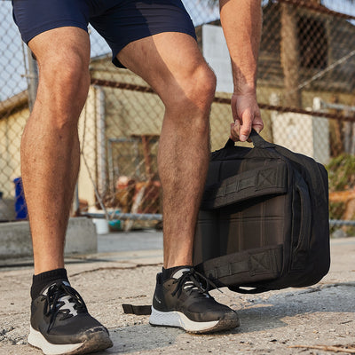 A person wearing black sneakers and navy shorts squats down to pick up a GORUCK Rucker 4.0 rucksack in an outdoor urban setting, framed by a chain-link fence and building in the background.