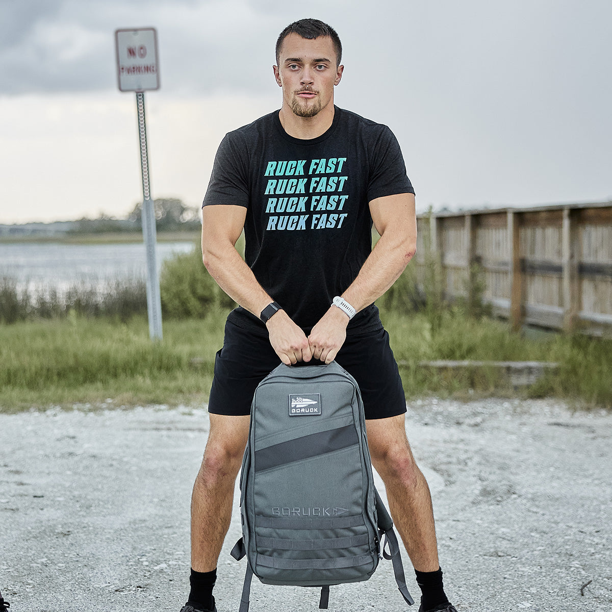 A man wearing a Ruck Fast T-shirt and shorts stands on a gravel path, holding his GORUCK Rucker 4.0 backpack by its straps. Positioned near the water and a wooden bridge, he captures the essence of rucking. A No Parking sign can be seen in the background.