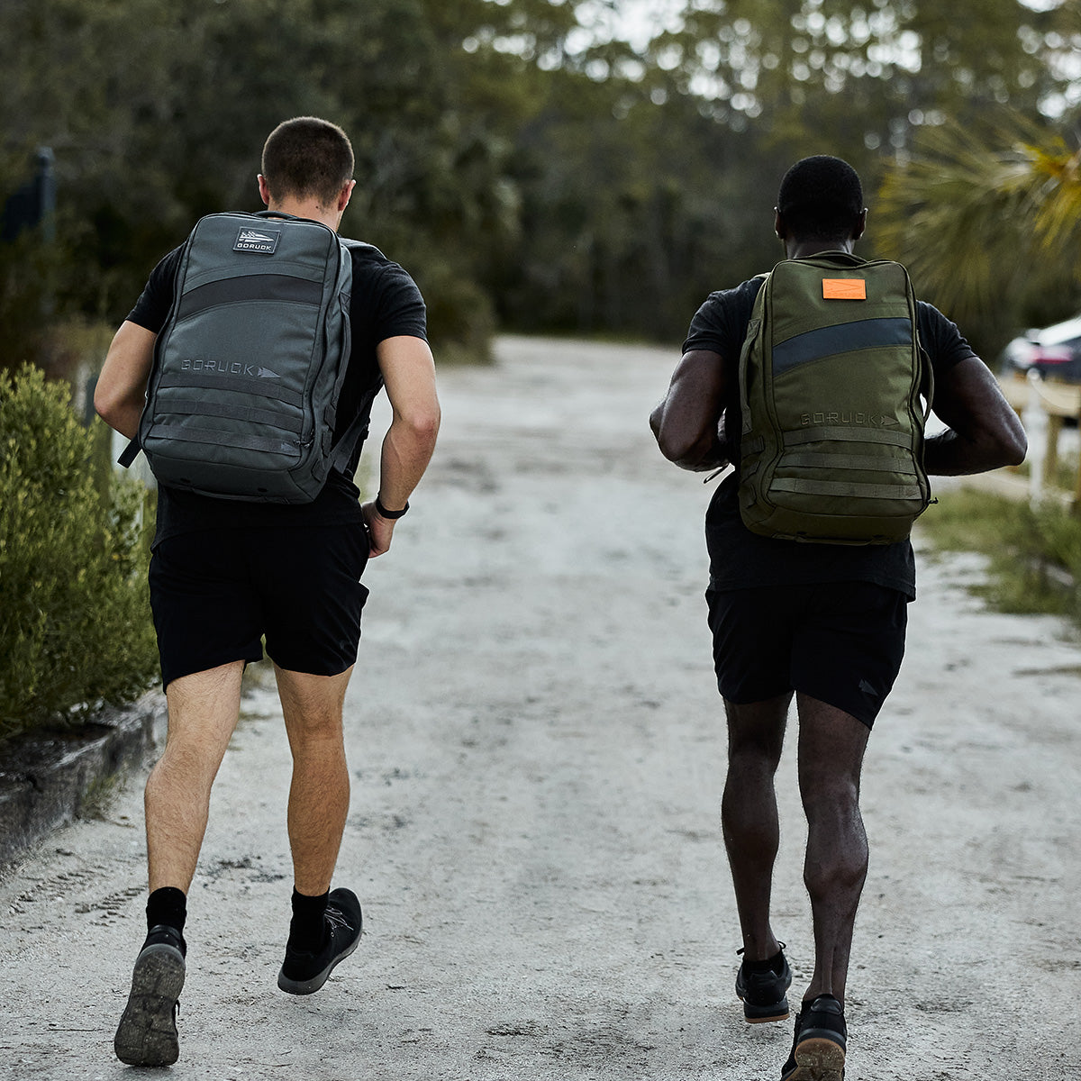 Two individuals walk along a dirt path enveloped in vibrant greenery, each carrying a Rucker 4.0 from GORUCK. One rucksack is gray while the other is green. Dressed in black attire, they partake in rucking as they move away from the camera.