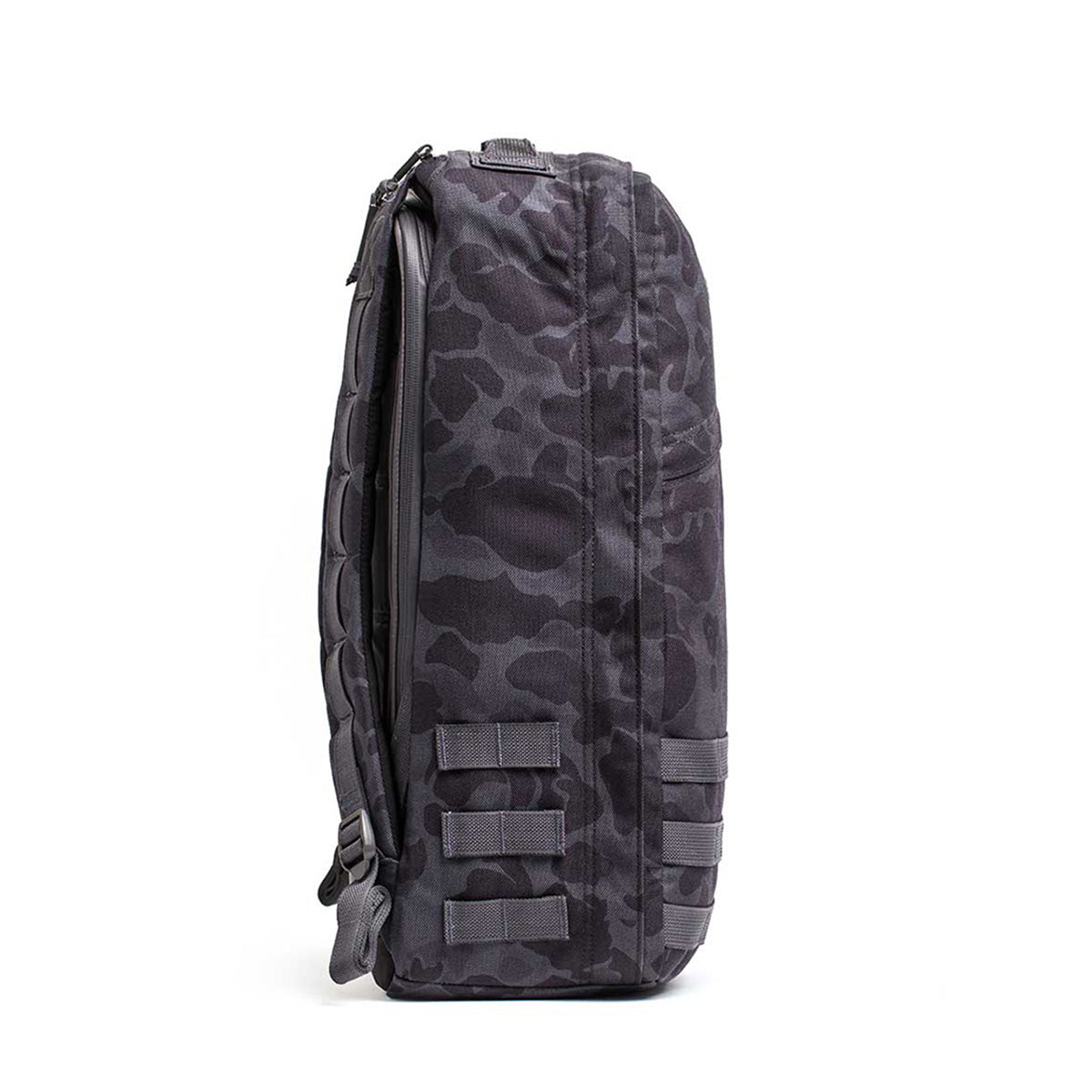 Side view of the GR1 USA - Cordura, a black camo-patterned backpack resembling a Special Forces rucksack, with multiple straps and zippers, ideal for carrying essentials during a GORUCK Challenge.