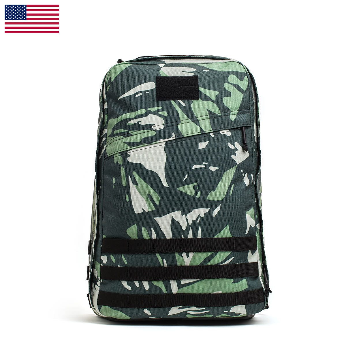 The GR1 USA x Gear Patrol, a limited-edition green camouflage backpack by GORUCK featuring black straps, is showcased against a white background. The distinctive variegated elephant ear camo pattern is prominent, with a small American flag accentuating the top left corner of the image.