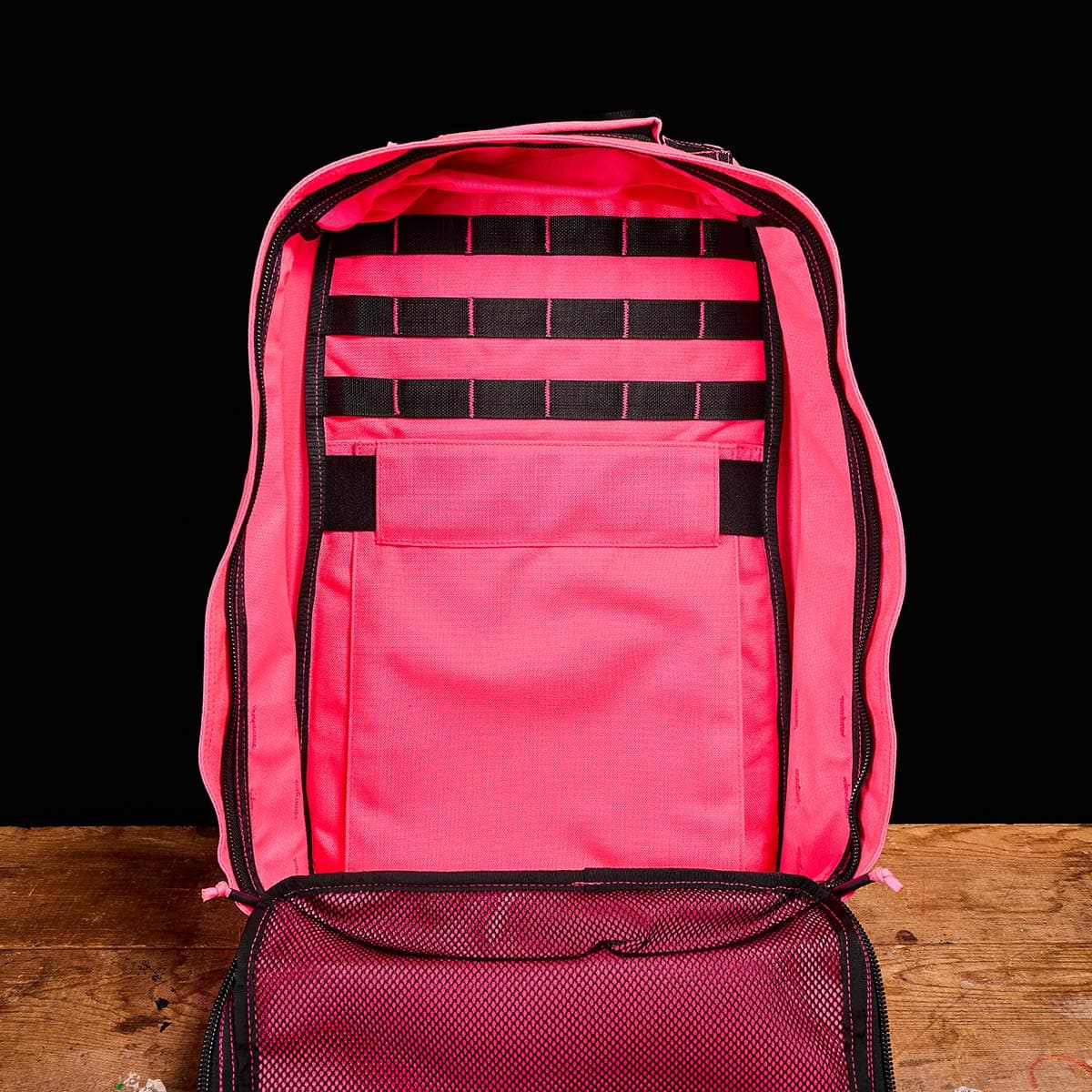 The GR1 USA - Hot Pink backpack by GORUCK is showcased against a black backdrop, displaying its roomy interior with mesh pockets and multiple horizontal straps for excellent organization. Crafted for performance and durability, the bag sits confidently on a wooden surface.