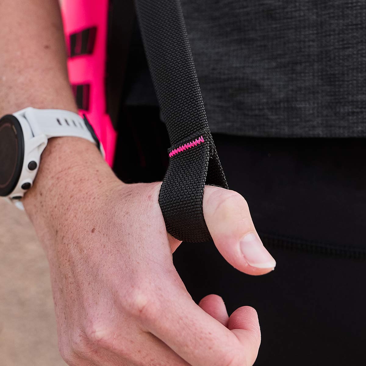 The image showcases a person wearing a smartwatch, illustrating resilience, while grasping the GR1 USA - Hot Pink strap from GORUCK, complete with a thumb loop. A small pink stitch on the strap subtly elevates its design. Clad in high-performance dark gray and black attire, this individual radiates confidence and strength.