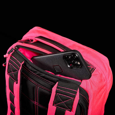 The GR1 USA - Hot Pink backpack by GORUCK, with its pink straps and black stitching, boldly contrasts against a solid black background as a sleek black smartphone peeks out of its main pocket, showcasing durability reminiscent of Special Forces equipment.