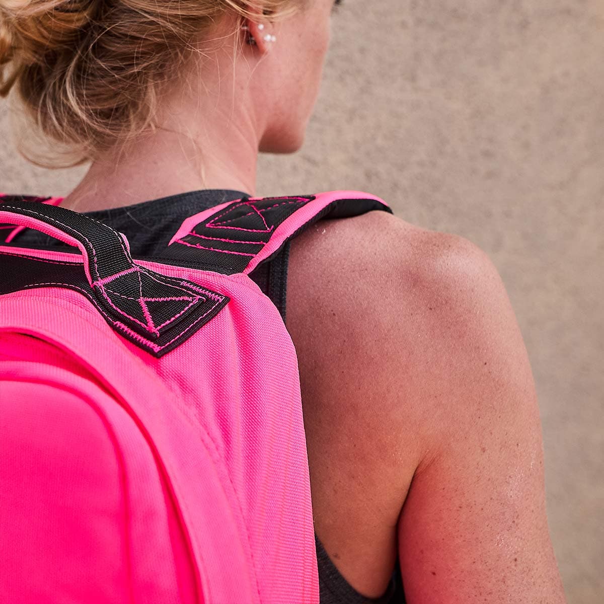 A person sporting the GR1 USA - Hot Pink backpack from GORUCK, its straps embellished with black geometric patterns, stands against a neutral-colored wall. Emulating the understated resilience of Special Forces, their short, light hair is pulled back as they confidently wear a sleeveless top.