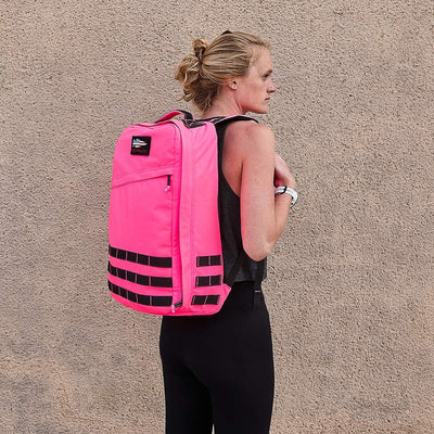 A person with blonde hair stands against a beige textured wall, exuding toughness in a black tank top and leggings. They're carrying the GR1 USA - Hot Pink backpack by GORUCK, featuring black straps and accents, ready for performance at any moment.