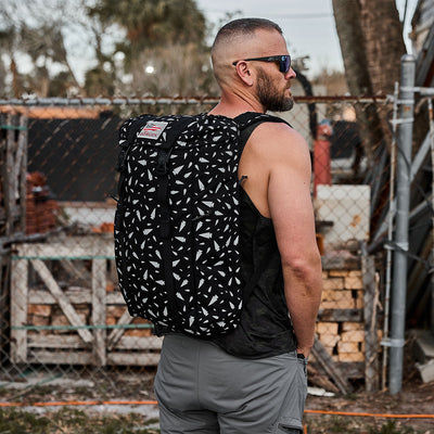 A man wearing sunglasses and a sleeveless top stands with his back to the camera, carrying a GORUCK M23 - Cordura - Spearhead Shrapnel, made from durable materials. He is in an outdoor setting near a fence with wood piles and trees in the background.