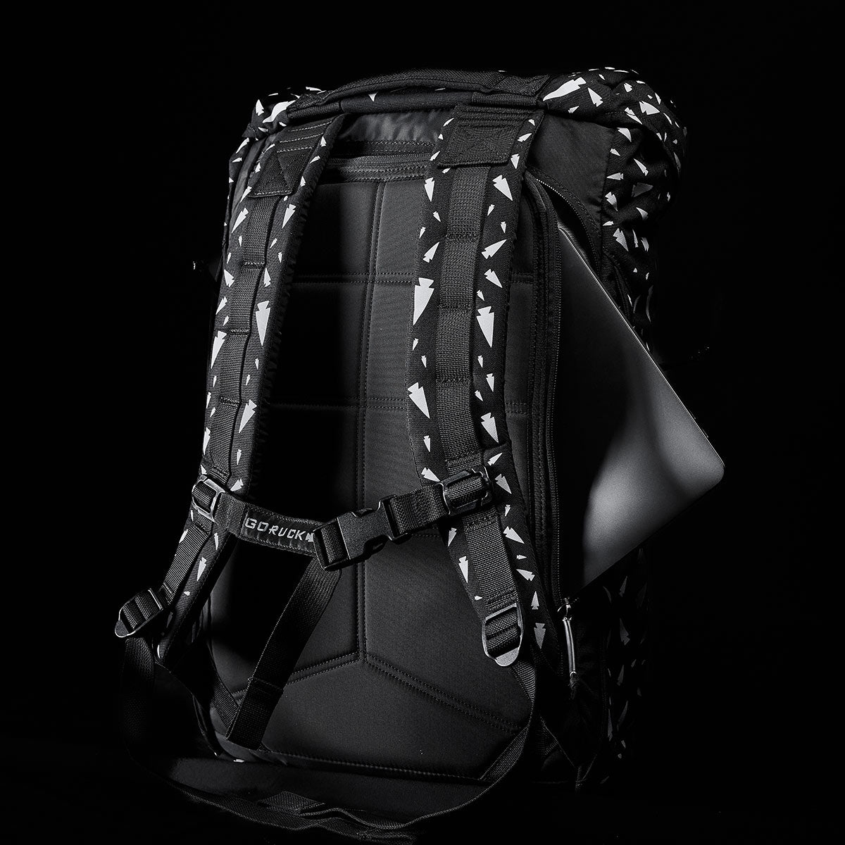 The M23 - Cordura - Spearhead Shrapnel by GORUCK is a contemporary rucksack made from durable materials, boasting a sleek black design adorned with geometric white patterns. It includes padded straps and a horizontal strap for extra support, with a laptop visible from the side pocket against its stark black backdrop.