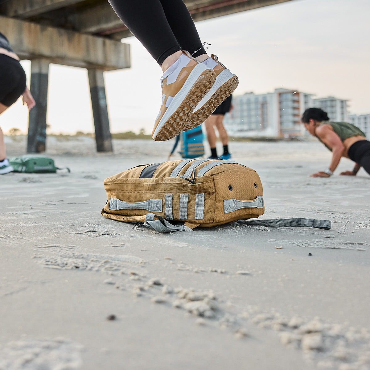 A person wearing the Women's Rough Runner - Speed Tan by GORUCK leaps over a yellow gym bag on a sandy beach. In the background, two others appear to be training for Rough Runner near a pier, with buildings silhouetted in the distance.