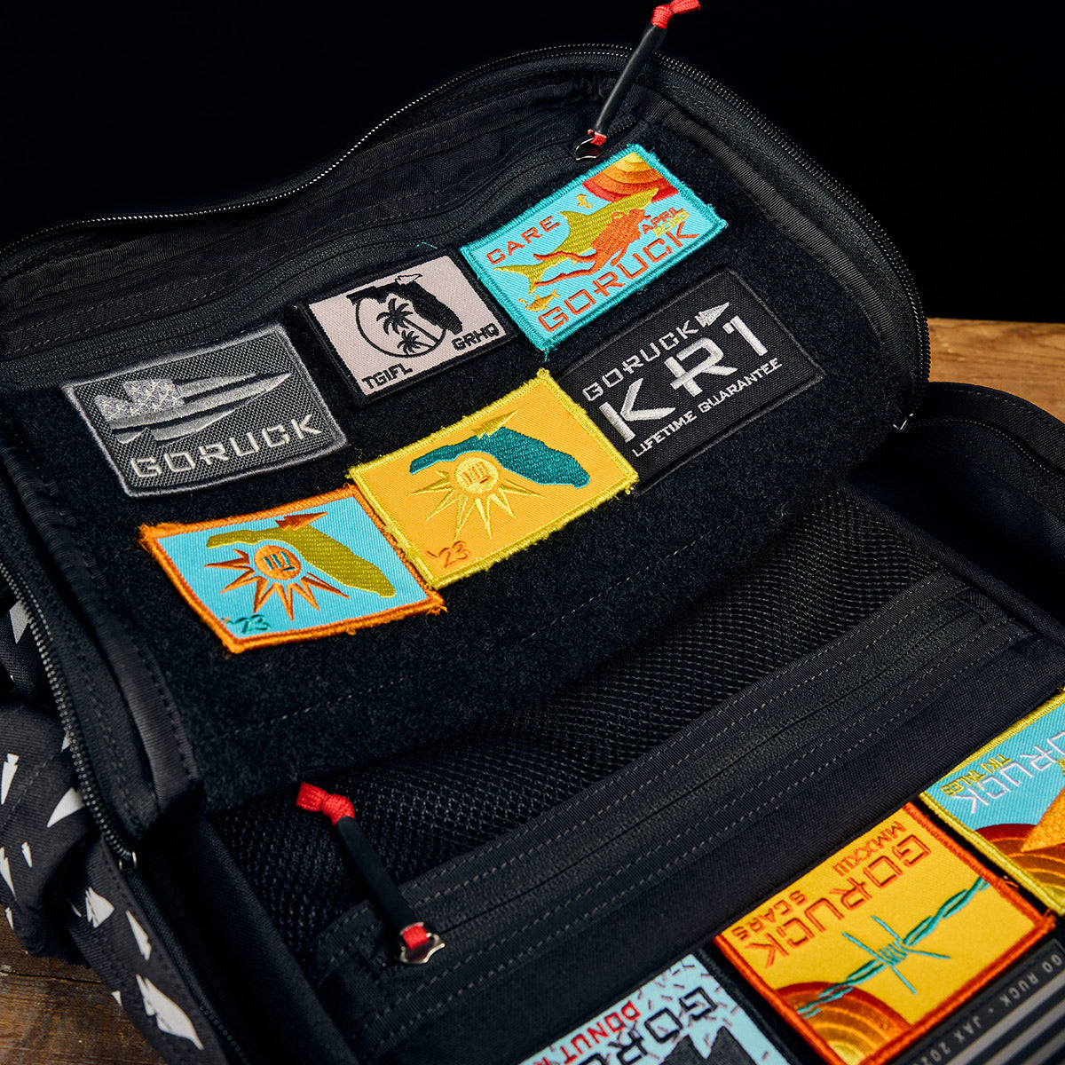 A black GORUCK KR1 - Spearhead Splatter kids' rucksack is displayed open on a wooden surface, featuring various colorful patches inside. The patches include abstract designs, logos, and text such as "GORUCK" and "LIFETIME GUARANTEE," making it perfect for carrying school supplies.