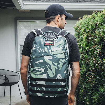 A man in a black cap and t-shirt stands outdoors, facing away. He's sporting the limited-edition GORUCK GR1 USA x Gear Patrol rucksack with variegated elephant ear camo. In the background, a potted plant, chair, and house exterior are visible.